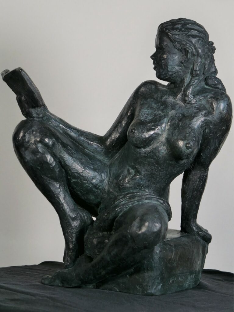 Image d’une sculpture en bronze représentant une figure féminine assise. La pose est décontractée et contemplative, avec un genou levé et l’autre replié sous elle. La figure tient un livre ouvert dans sa main gauche, le regardant avec attention, évoquant une scène de lecture tranquille. Ses cheveux sont coiffés en arrière dans un style désinvolte. La sculpture est placée sur une surface plane et claire, avec un fond légèrement flou qui semble être l’intérieur d’un bâtiment avec des éléments de décor. Une plante aux feuilles vertes est visible en arrière-plan sur la droite, ajoutant une touche de vie à la composition.
Sculpteur Jean-Pierre Picheny, influences Rodin, Claudel, réalise aussi des lampes et exécute des moulages sur corps