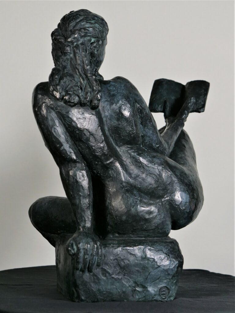 Image d’une sculpture en bronze représentant une figure féminine assise. La pose est décontractée et contemplative, avec un genou levé et l’autre replié sous elle. La figure tient un livre ouvert dans sa main gauche, le regardant avec attention, évoquant une scène de lecture tranquille. Ses cheveux sont coiffés en arrière dans un style désinvolte. La sculpture est placée sur une surface plane et claire, avec un fond légèrement flou qui semble être l’intérieur d’un bâtiment avec des éléments de décor. Une plante aux feuilles vertes est visible en arrière-plan sur la droite, ajoutant une touche de vie à la composition.
Sculpteur Jean-Pierre Picheny, influences Rodin, Claudel, réalise aussi des lampes et exécute des moulages sur corps